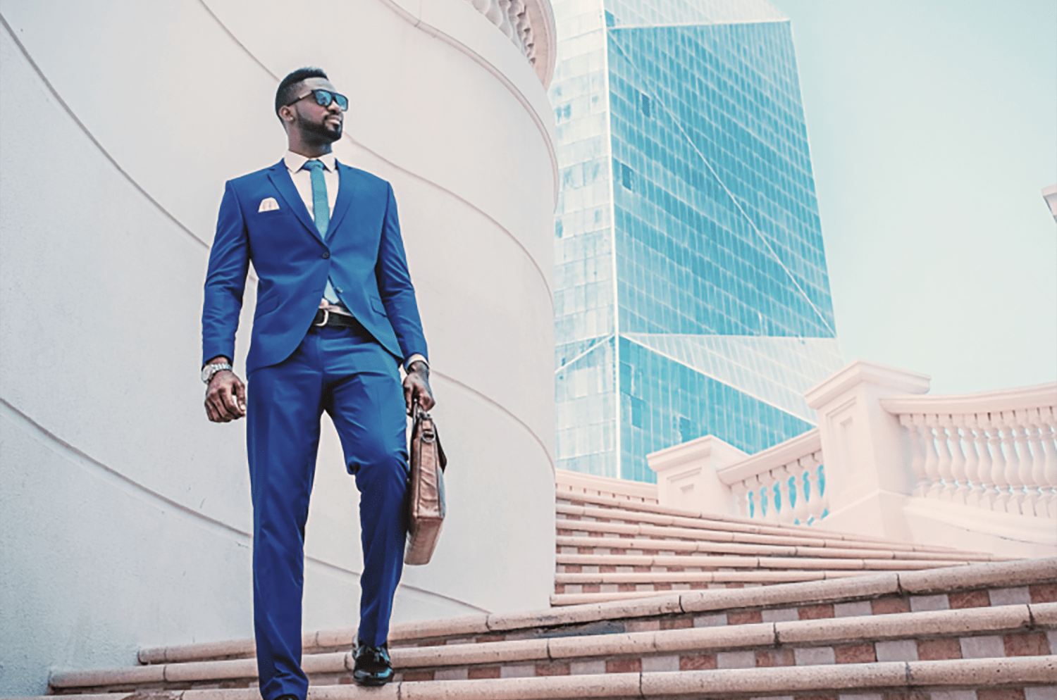An african man in a blue suit and tie walking down a stairs in front of a skyscraper